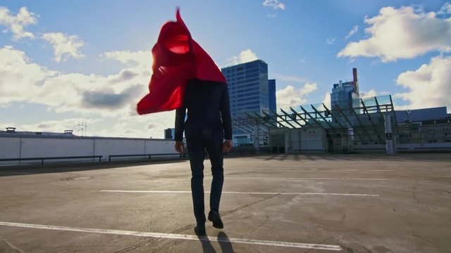 Businessman Superhero WIth Red Cape Blowing in the Wind Walks on the Roof of a Skyscraper Ready to Make Business Transactions and Save the Day. Following Back View Shot.