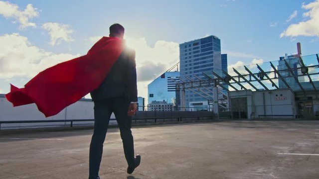 Businessman Superhero With Red Cape Blowing in the Wind Walks on the Roof of a Skyscraper Ready to Save the Day. In the Background Modern City Center. Following Back View Shot. 