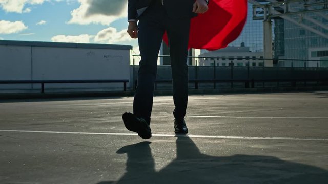 Businessman Superhero With Red Cape Blowing in the Wind Walks on the Roof of a Skyscraper Ready to Save the Day. In the Background Modern City Center. Elevating Shot In Slow Motion. 