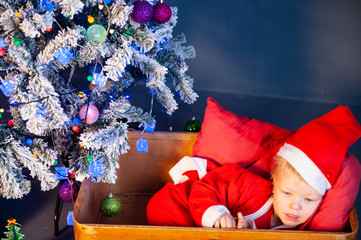 Happy child in red suit settles down to sleep in wooden box under the Christmas tree in...