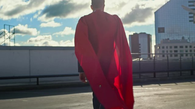 Businessman Superman WIth Red Cape Blowing in the Wind Walks on the Roof of a Skyscraper, Looking into the Sunset, Ready to Save the Day. Following Back View Slow Motion Shot.