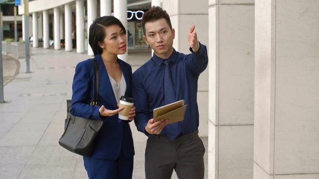 Medium shot of young Asian businessman holding tablet, showing something in it to Asian businesswoman standing nearby and then comparing it with objects which surround them