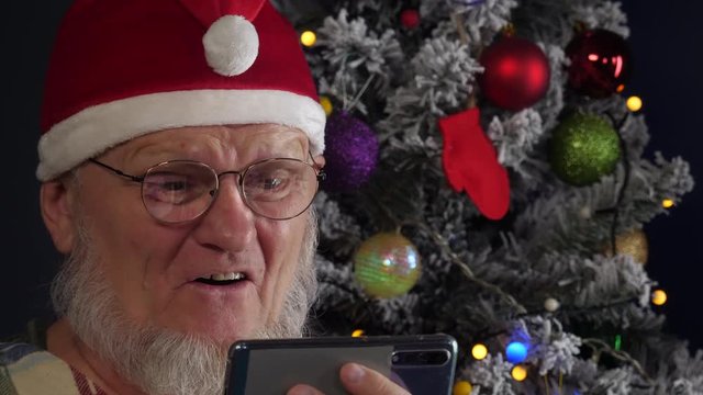 Happy Elderly man in Santa Claus hat is chatting with friends using smartphone and actively expresses his emotions against background of Christmas tree in garlands, green UFO balls, proton purple toy