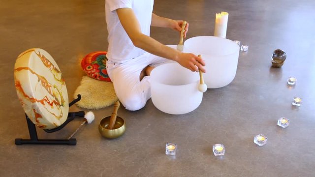 Man playing crystal bowls healing music. Fixed angle scene of a young man dressed in white, sitting in indian surrounded by instruments and playing his two crystal bowls as part of a meditative concer