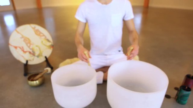 Man playing crystal bowls healing music. Fixed angle scene of a young man dressed in white, sitting in indian surrounded by instruments and playing his two crystal bowls as part of a meditative concer