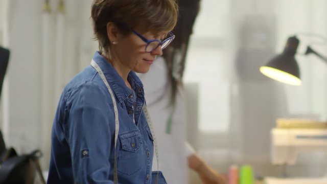 Tilt down of middle aged Caucasian woman in eyeglasses drawing with tailors chalk on fabric and then taking measuring tape while making clothes in sewing studio