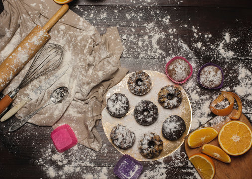 A photo of tasty cakes sprinkled with sweet powder and chocolate on a wooden brown table. Oranges and instruments  for prepare cakes. Composition with cupcakes on the table