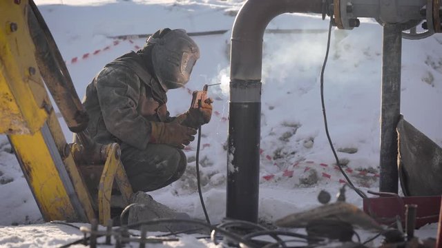 Worker produces pipe welding on the street. In winter, at low temperatures, it is difficult to mount the pipe wire.