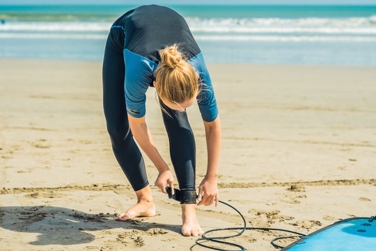 Summer time and active rest concept. Young surfer woman beginner fastens leash across leg, going to surf on big barral waves on ocean, dresseed in boardies, poses aganist sky on hot sunny day