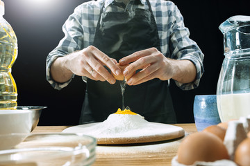 Professional male cook sprinkles dough with flour, preapares or bakes bread or pasta at kitchen table, has dirty uniform, isolated over black chalk background. Baking concept