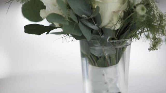 Beautiful bouquet of white flowers with big green leaves in glass vase put on white table in room with grey walls.