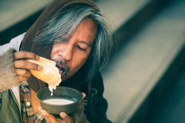 Male Beggar, Homeless man on the stair of the walkway street in the city eating bread and milk form...