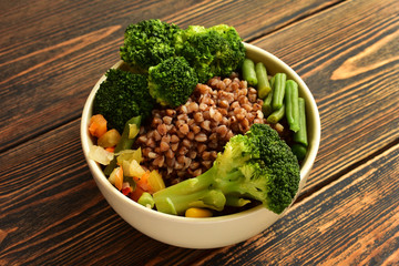 A bowl of delicious buckwheat porridge with vegetables and rye bread located on a dark wooden background.