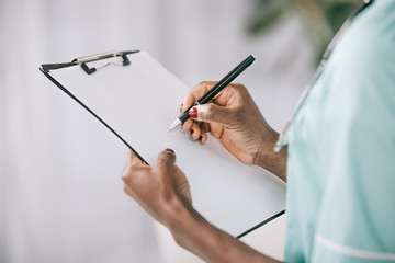 cropped view of african american nurse writing in clipboard with paper