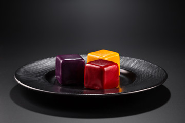 three miniature cakes in the shape of a cube of different colors on a black plate.