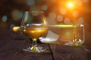 Glass of whiskey or brandy and empty bottles on wooden table on bright glowing background