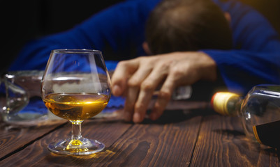 Male alcoholic with glass of whiskey and bottle sleeping on table at night.