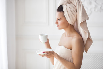 Attractive young woman drinking coffee or tea after shower. Beautiful girl in the towels at morning. Lady after bathing. Close up portrait of female indoors