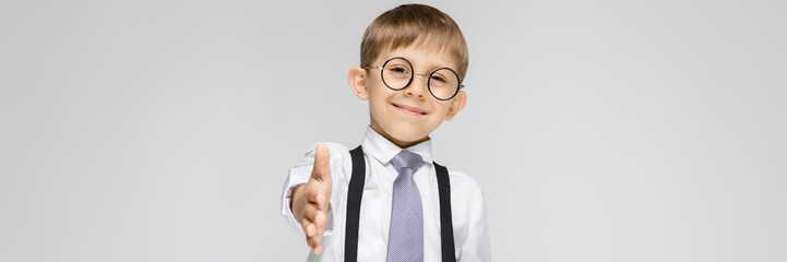 A charming boy in a white shirt, suspenders, a tie and light jeans stands on a gray background. The boy stretches out his hand forward