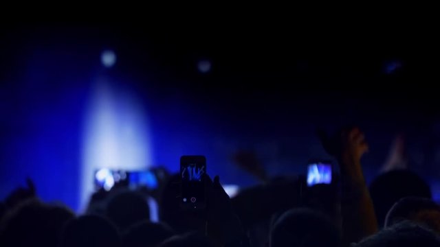 People crowd partying at music concert, jumping and clap their hands against strobing flashing lights, dancing, taking photos and recording video with smart phones