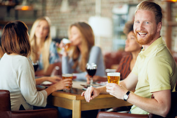 Man looking at camera and drinking beer while sitting in restaurant. In background friends chatting.
