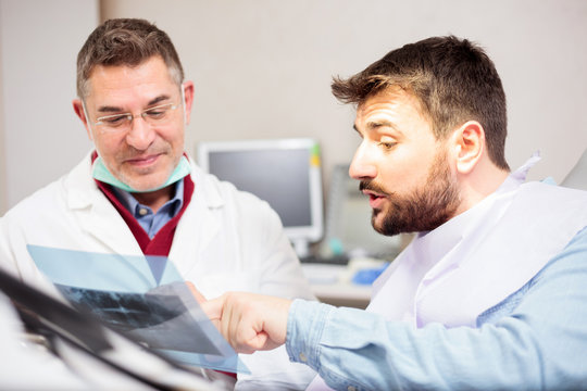 Mature male dentist showing x-ray image to a young male patient during the exam in dental clinic. Health care and medicine concept.