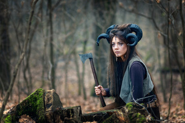 Beautiful young woman warrior with blue eyes wearing ram horns and specific makeup holding ax hiding behind stumps in forest, stalking and ready to attack or defend her land.