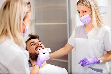 Two young female dentists working in dental clinic. Whitening male patient's teeth and using tone chart to mach color. Health care and medicine concept.