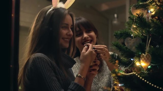 Girlfriends decorate a christmas tree with joy and in festive mood. Two girls Laughing while dressing up christmas tree. Sisters together fill the Christmas tree with toys, preparing for holidays