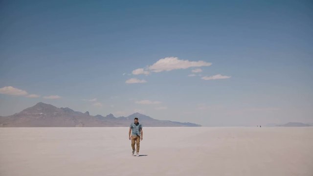Tired and exhausted young man walking towards camera looking around at epic hot sunny salt desert lake in Utah USA.