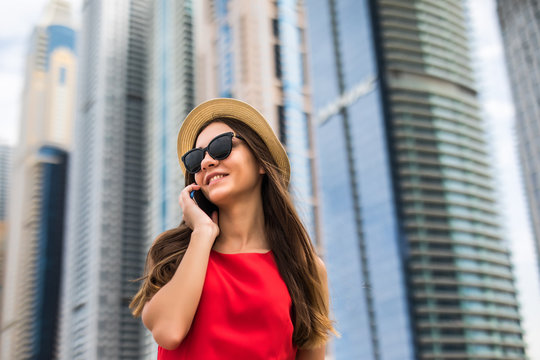 Portrait of young smile woman in red dress, sunglasses and summer hat talking on the phone on downtown skycrapers background