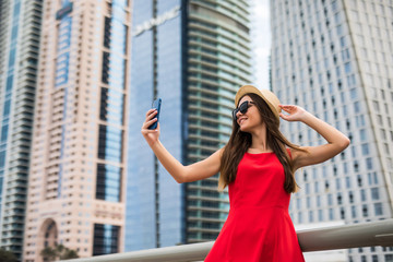 Portrait of young smile woman in red dress, sunglasses and summer hat take selfie on the phone on downtown skycrapers background