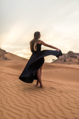 Young pretty woman in black dress dancing in sandy desert at sunset