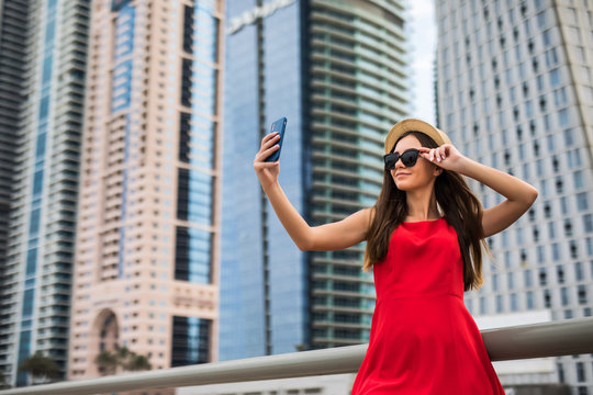 Portrait of young smile woman in red dress, sunglasses and summer hat make selfie or video call on the phone on downtown skycrapers background