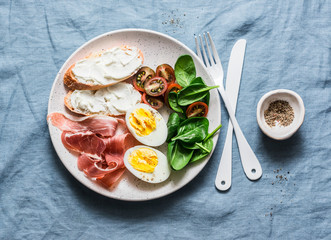 Cream cheese toast, boiled egg, prosciutto, spinach, tomatoes - delicious healthy breakfast or snack on blue background, top view
