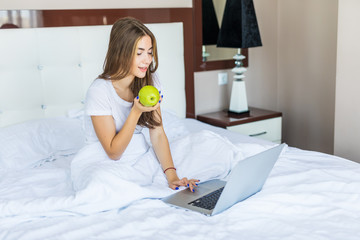 Portrait of girl sits in bed early in the morning, eats an apple and smiles, with a laptop