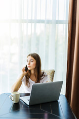 Smiling woman using mobile phone while holding cup of coffee and sitting with laptop computer at home