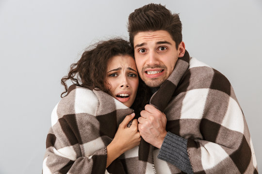 Photo of frozen couple man and woman wrapped in blanket trembling and hugging together, isolated over gray background