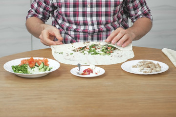 Man is cooking shawarma on the kitchen table at home. Pita, vegetables and green onion with sauce and mayonnaise. Close-up hands.