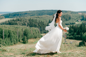 Fototapeta premium Happy brunette bride in beautiful white dress posing with boquet of flowers on top of mountain with fabulous landscape view. Bridal outdoor photosession. Cheerful brunette bride lifestyle portrait.