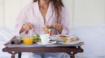 Obraz na płótnie Canvas Making breakfast in bed / pretty smiling brunette woman holding a tray with healthy food while sitting on the bed