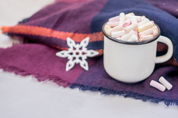 Obraz na płótnie Canvas Winter cozy concept. Coffee with marshmallows and decorative shiny snowflake in white enameled metal cup in purple warm wool scarf. Warm weekend in cold weather.