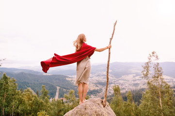 Unknown female in dress and red cape standing on stone at top of mountaing. Barefoot blonde woman...