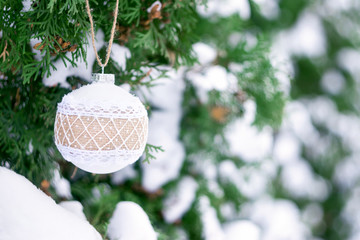 Christmas winter holiday greeting card.  White rustic Christmas ornament ball with burlap on green christmas trees with snow. Festive outdoor background. Xmas composition decoration