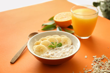 Tasty oatmeal with banana slices in bowl and glass of orange juice on color table