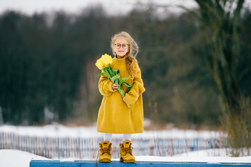 Funny little girl in big adudlt oversized yellow woolen shirt and yellow boots holding bpuquet of yellow tulips outdoor. Kids fashion. Adorable child with braid in glasses lifestyle winter portrait.
