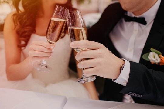 Cropped image of bride and groom clink glasses of champagne during their wedding day, dressed in white dress and black suit, celebrate creating family. Celebration, festive event concept. Newlyweds