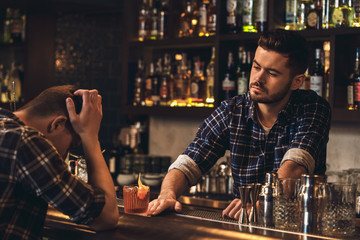 Young bartender standing at bar counter giving cocktail to sad client
