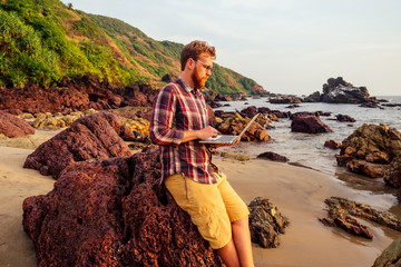 male freelancer bearded glasses hipster with a laptop remote work on the beach on the...