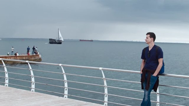 4K A handsom man is standing on a pier. There could be seen the wavecutter and a ship on the background